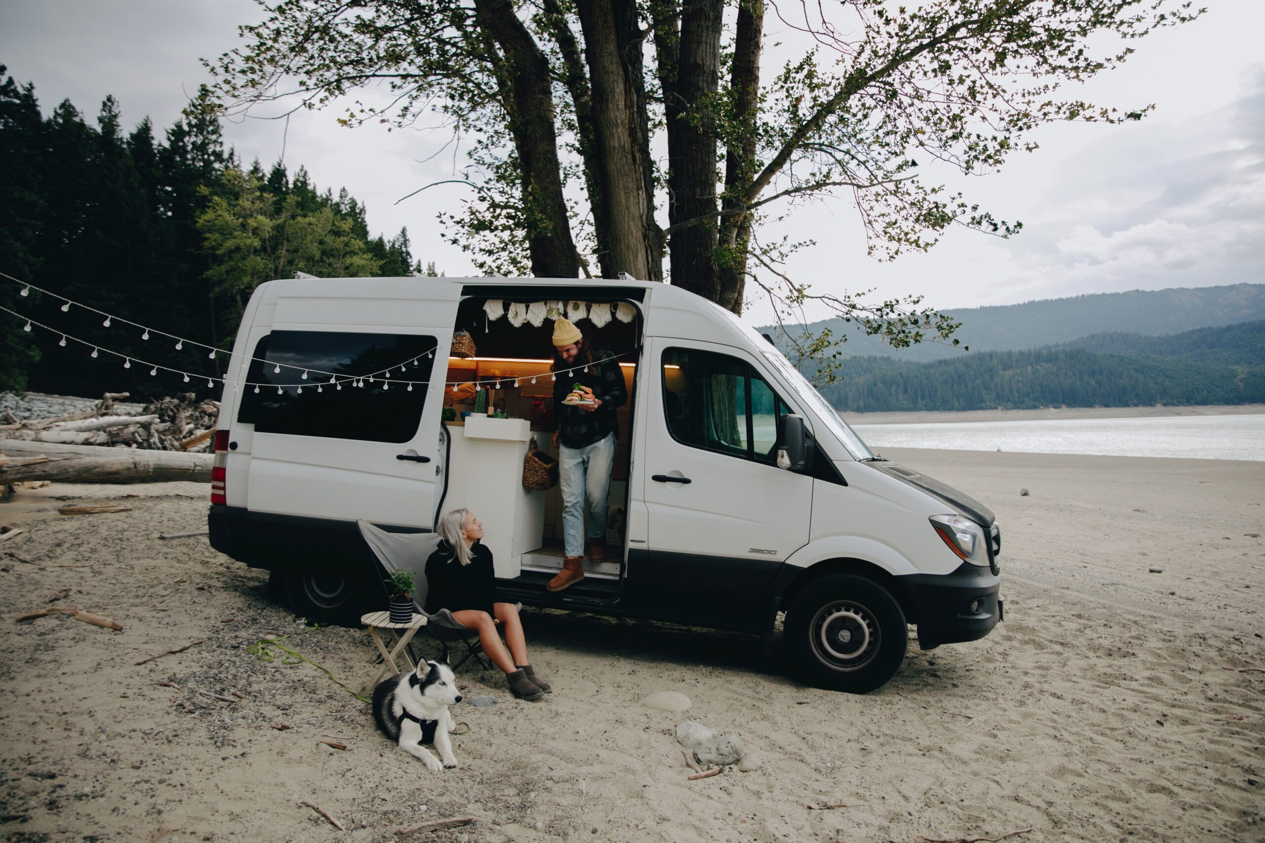 Couple with a dog relaxing outside an RV parked near a lake, representing MarCELL RV and pet monitoring for safe travel.